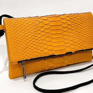 May include: A yellow snakeskin clutch bag with a black adjustable strap. The bag has a zipper closure and a flap.