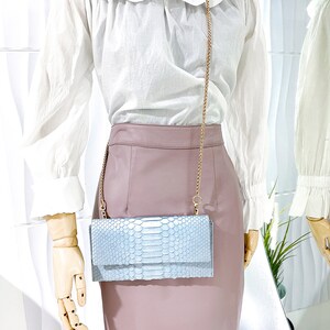 May include: A light blue snakeskin clutch bag with a gold chain strap. The bag is worn by a mannequin wearing a white blouse and a dusty rose skirt.