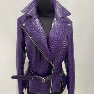 May include: A purple snakeskin-textured jacket with a notched collar, asymmetrical zipper closure, and a belted waist. The jacket features long sleeves, silver zippers, and a matching belt. The jacket is displayed on a black mannequin.