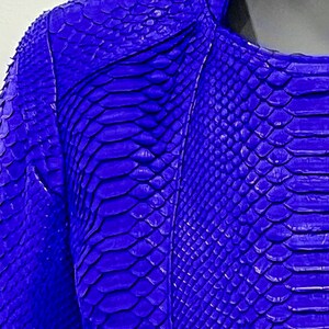 K&ouml;nnte beinhalten: Eine leuchtend blaue Schlangenlederjacke mit Rei&szlig;verschluss. Die Jacke hat ein strukturiertes, schuppiges Muster.
