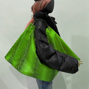 以下が含まれることがあります： シルバーの金具が付いた、鮮やかな緑色のヘビ柄の大きなハンドバッグ。黒いパファージャケットと青いジーンズを着たマネキンが持っています。