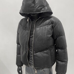 Op de afbeelding: Zwarte pufferjack met capuchon, met een slangenleerpatroon. De jas heeft een volledige ritssluiting en wordt gedragen over een donker shirt en een blauwe jeans. De handen van de mannequin zijn zwart.