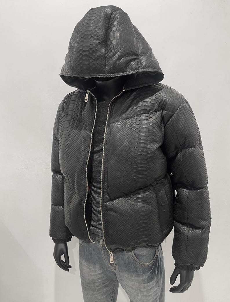 Op de afbeelding: Zwarte pufferjack met capuchon, met een slangenleerpatroon. De jas heeft een volledige ritssluiting en wordt gedragen over een donker shirt en een blauwe jeans. De handen van de mannequin zijn zwart.