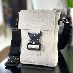 May include: A white snakeskin crossbody bag with a black strap and a black buckle closure.