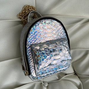 May include: A silver iridescent snakeskin backpack with a gold chain strap and a zipped front pocket.