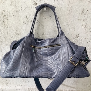 May include: A dark blue leather duffel bag with a snakeskin pattern. The bag has a top handle and a long adjustable shoulder strap.