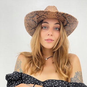May include: A woman wearing a brown snakeskin cowboy hat with a black and white polka dot off-the-shoulder blouse. She is holding a black smartphone in her hands.