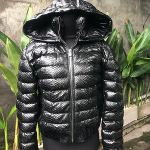 May include: A black puffer jacket with a hood made of snakeskin. The jacket has a zip fastening and a quilted design.