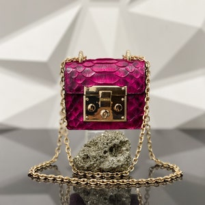 May include: A small, pink, snakeskin handbag with a gold chain strap and a gold lock. The bag is sitting on a rock.