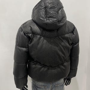 Op de afbeelding: Zwarte pufferjack met capuchon, met een textuur die op slangenleer lijkt. De jas heeft een trekkoord in de taille en wordt op een mannequin getoond. De jas is donker van kleur.