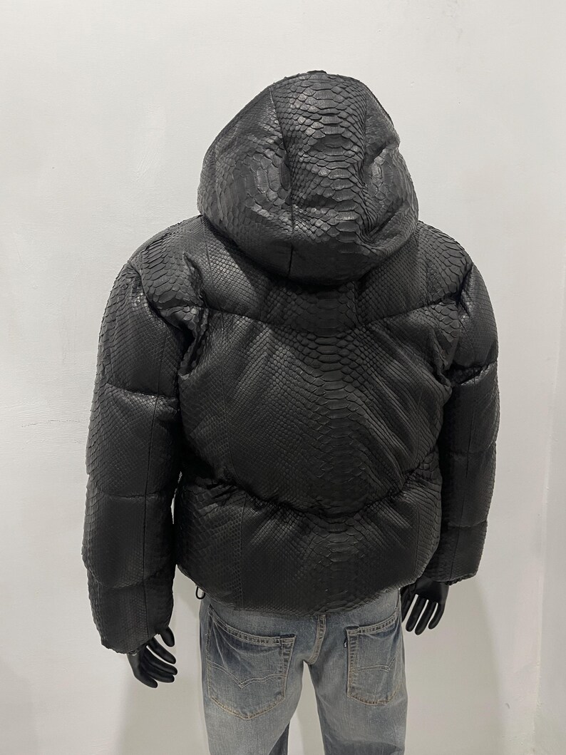 Op de afbeelding: Zwarte pufferjack met capuchon, met een textuur die op slangenleer lijkt. De jas heeft een trekkoord in de taille en wordt op een mannequin getoond. De jas is donker van kleur.