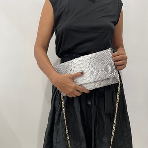 May include: A silver and white clutch with a snakeskin pattern and a gold chain strap. The clutch is rectangular and folded over. The person is wearing a black dress.