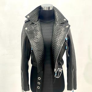 May include: Black leather biker jacket with a snakeskin effect. The jacket has a notched collar, silver-tone metal hardware, and a waist belt. Zips are visible on the sleeves. The jacket is displayed on a mannequin.