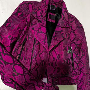 May include: A pink and black snakeskin leather jacket with a zip fastening and a belt. The jacket has a black lining and a label that says "SUGAR" on the inside.