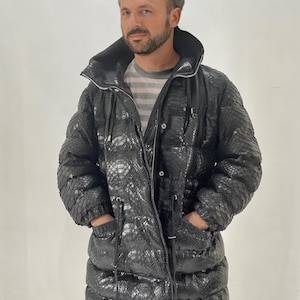 以下が含まれることがあります： 黒いヘビ柄のロングパファージャケット。 ジャケットはハイカラー、ジッパー開閉、ポケット付き。 ストライプのシャツの上にジャケットを着ています。