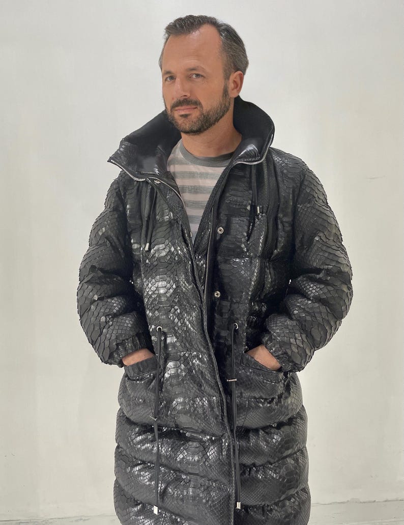 May include: A black, long puffer coat with a snakeskin pattern. The coat has a high collar, a zipper closure, and pockets. The coat is worn over a striped shirt.