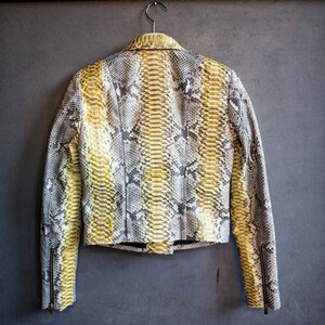 以下が含まれることがあります： イエロー、グレー、白の色合いのヘビ柄のクロップドジャケット。 ジャケットは襟付き、袖口にジッパー付きの長袖、テーラードフィットです。 グレーの背景に金属製のハンガーに掛けられています。