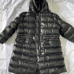 May include: Black puffer coat with a hood. The coat has a textured, snake-like pattern and a drawstring waist. It features a zipper closure and is designed for cold weather. The coat is laid out on a white surface.