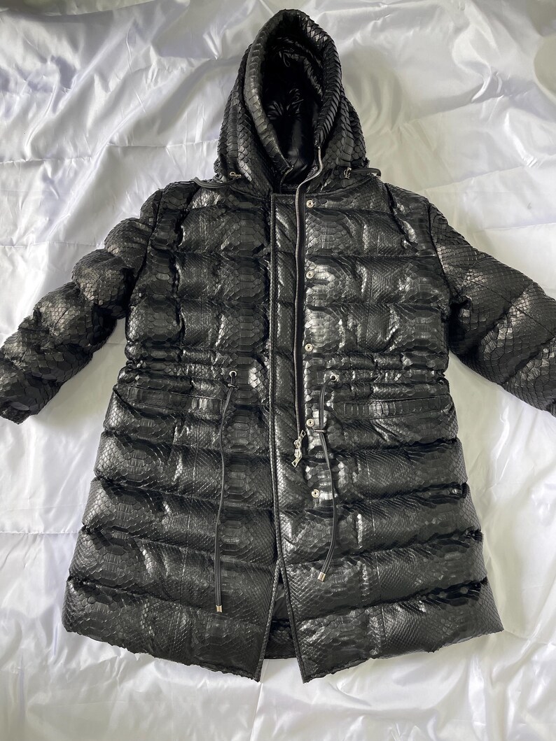 May include: Black puffer coat with a hood. The coat has a textured, snake-like pattern and a drawstring waist. It features a zipper closure and is designed for cold weather. The coat is laid out on a white surface.