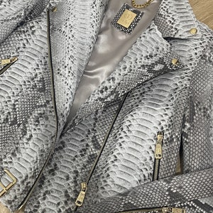 May include: A gray snakeskin leather jacket with a gold zipper and a gold chain. The jacket has a satin lining and a label that says "COLLI" on it.