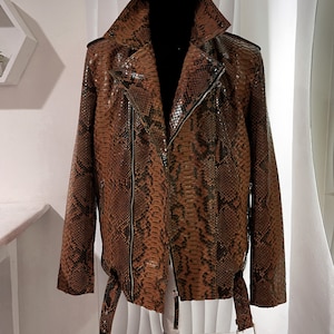 以下が含まれることがあります： ジッパー留めとベルトが付いた茶色のヘビ革ジャケット。ジャケットはスタンドカラーと長袖です。