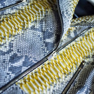 以下が含まれることがあります： グレーとイエローの色合いのヘビ革模様のジャケットのクローズアップ。 ジャケットはシルバーのジッパーとテクスチャ加工された表面が特徴で、デザインの複雑なディテールを示しています。 全体的なテーマはファッションとアパレルです。