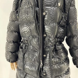 May include: A long, black puffer coat with a snakeskin pattern. The coat features a hood, a zipper closure, and pockets. The coat has a drawstring waist and is made of a textured material. The coat is designed for cold weather.