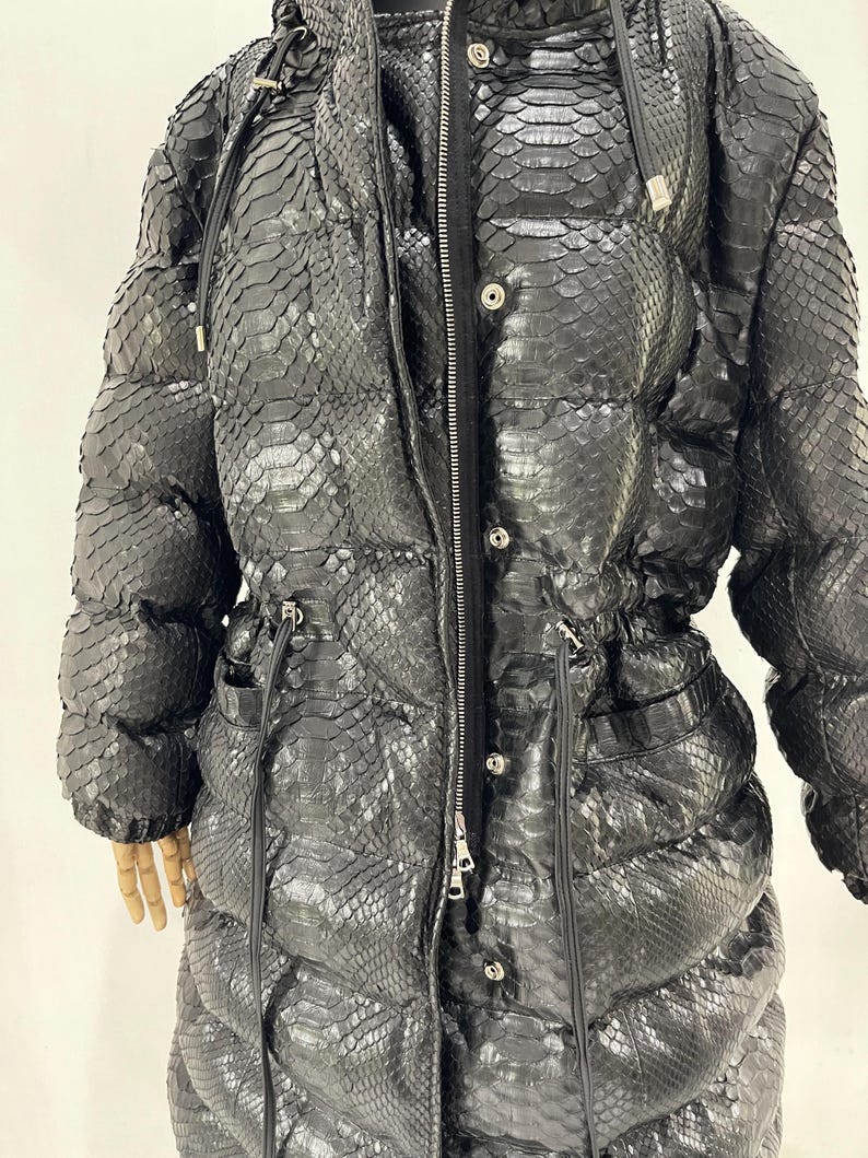 May include: A long, black puffer coat with a snakeskin pattern. The coat features a hood, a zipper closure, and pockets. The coat has a drawstring waist and is made of a textured material. The coat is designed for cold weather.