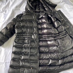 May include: Black puffer jacket with a hood. The jacket has a snakeskin-like texture on the outer layer and a shiny, quilted interior. A drawstring cinches the waist. The jacket is open, revealing the interior lining.