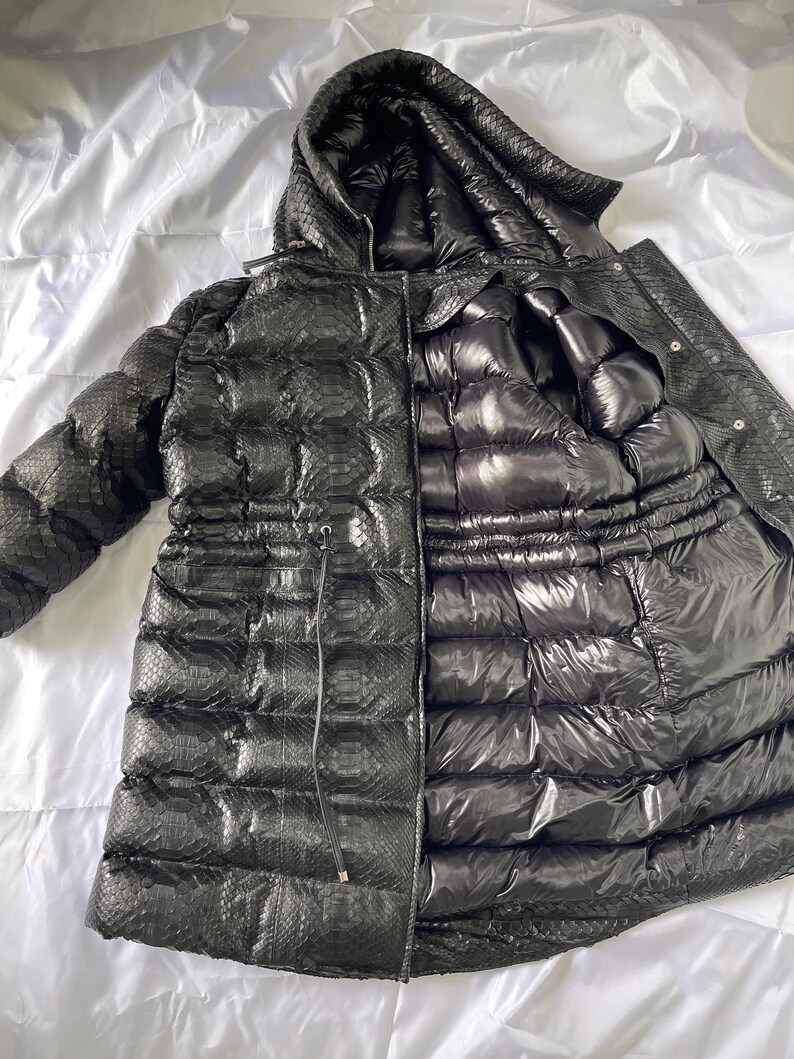 May include: Black puffer jacket with a hood. The jacket has a snakeskin-like texture on the outer layer and a shiny, quilted interior. A drawstring cinches the waist. The jacket is open, revealing the interior lining.
