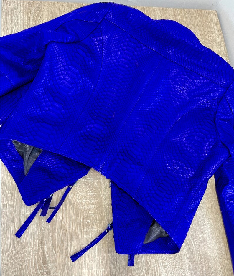 K&ouml;nnte beinhalten: Eine blaue Schlangenlederjacke im Cropped-Stil mit Rei&szlig;verschl&uuml;ssen. Die Jacke hat eine strukturierte Oberfl&auml;che und ein einzigartiges Design.