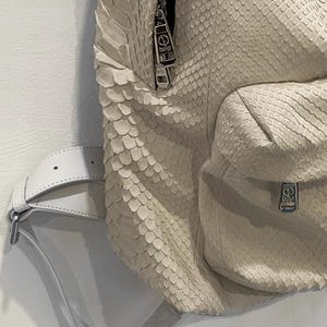 Op de afbeelding: Een witte slangleren rugzak met zilveren hardware en een witte leren riem. De rugzak heeft een ritssluiting en een klein vakje aan de voorkant.