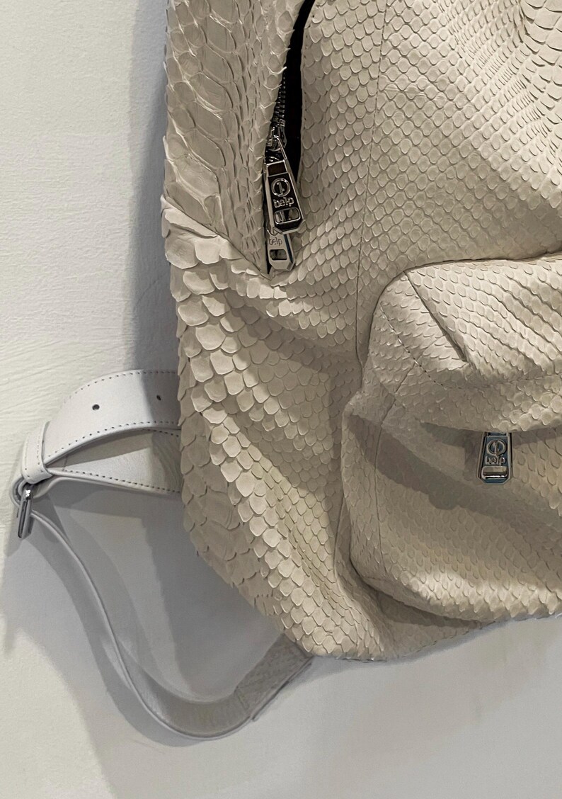 Op de afbeelding: Een witte slangleren rugzak met zilveren hardware en een witte leren riem. De rugzak heeft een ritssluiting en een klein vakje aan de voorkant.