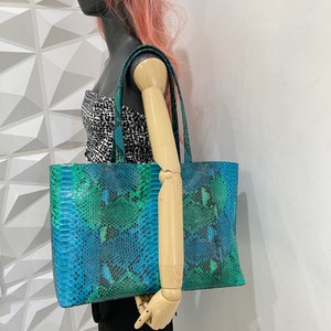 以下が含まれることがあります： 青と緑のヘビ柄の大きなトートバッグ。バッグにはトップハンドルが2つ付いています。