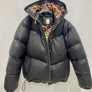 May include: Black puffer jacket with a snakeskin texture and leopard print lining. The jacket features a hood and a zip closure. It is suspended from a metal hanger.