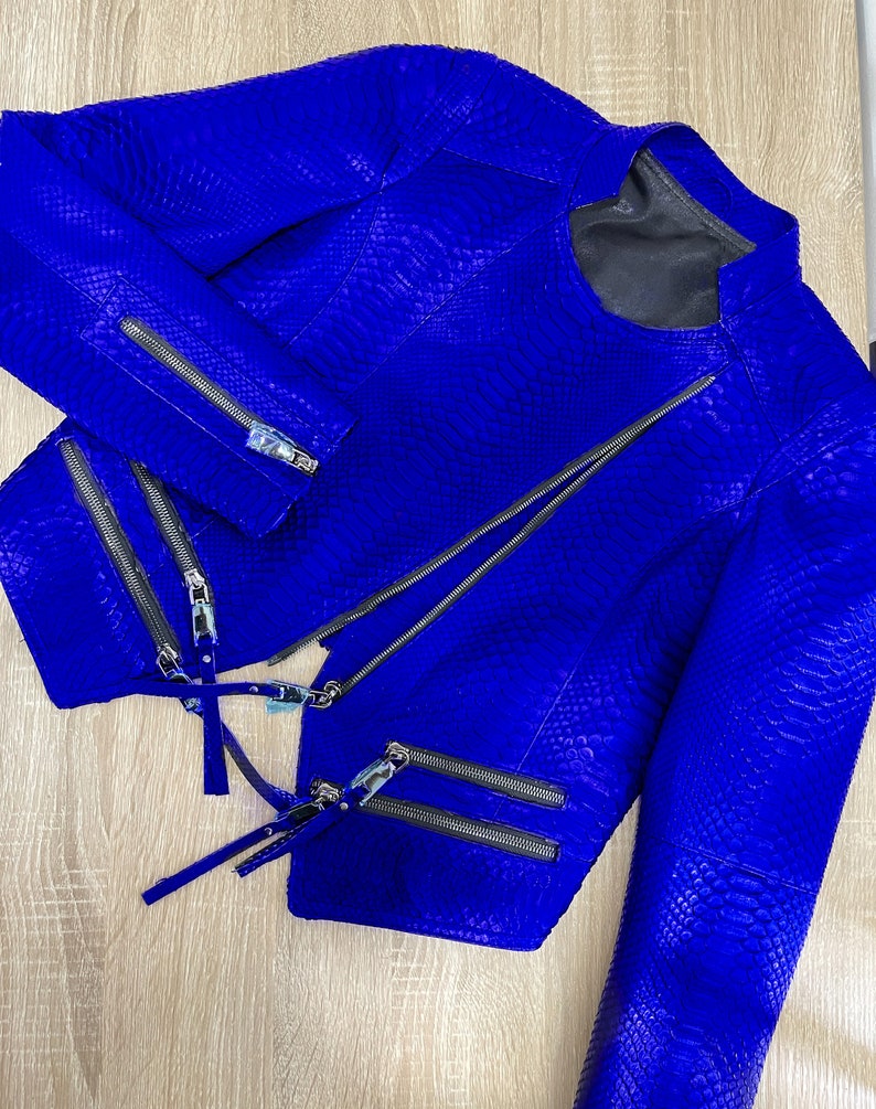 K&ouml;nnte beinhalten: Eine leuchtend blaue Schlangenlederjacke mit mehreren Rei&szlig;verschl&uuml;ssen. Die Jacke hat einen Stehkragen und lange &Auml;rmel.