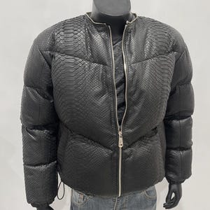 Op de afbeelding: Zwarte pufferjack met een slangenprint. De jas heeft een ritssluiting aan de voorkant en een ronde kraag. De jas wordt op een mannequin getoond. De jas wordt gecombineerd met een blauwe jeans.