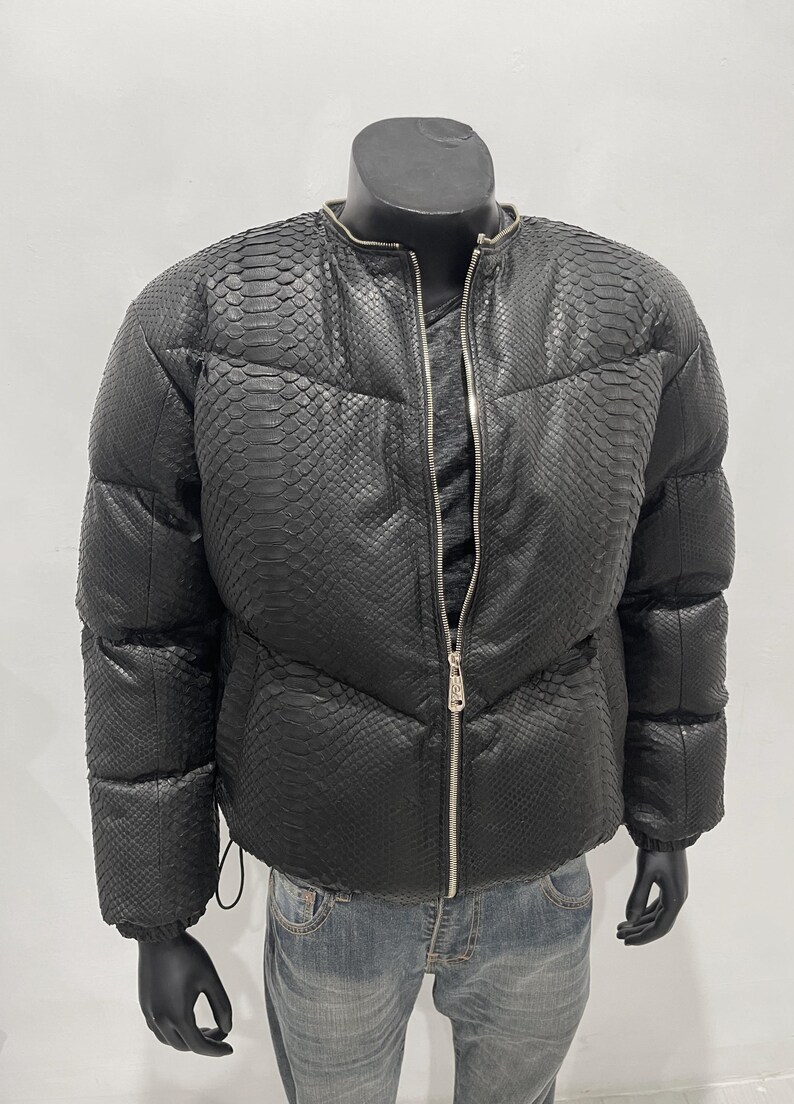 Op de afbeelding: Zwarte pufferjack met een slangenprint. De jas heeft een ritssluiting aan de voorkant en een ronde kraag. De jas wordt op een mannequin getoond. De jas wordt gecombineerd met een blauwe jeans.