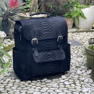 May include: Black leather backpack with a snakeskin pattern and two buckle closures. The backpack has a large front pocket and a zippered top closure.