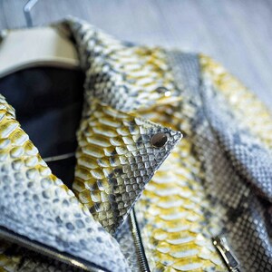 以下が含まれることがあります： ヘビ革で作られたスタイリッシュなジャケットのクローズアップ。 ジャケットは、黄色、グレー、白の鱗の混合が特徴で、襟には黒い裏地が見えます。 シルバーのメタルボタンとジッパーがディテールを追加しています。