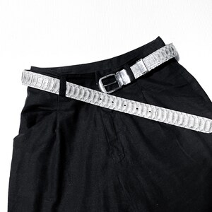 May include: A light gray belt with a reptile skin pattern is draped over a pair of black pants. The belt has a silver buckle and a series of evenly spaced holes for adjustment. The belt is made of a textured material.