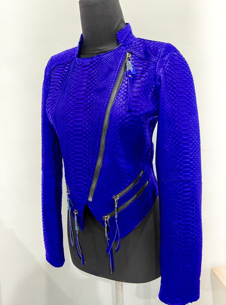 K&ouml;nnte beinhalten: Eine blaue Lederjacke mit Schlangenhautmuster und mehreren Rei&szlig;verschl&uuml;ssen. Die Jacke hat einen Stehkragen und lange &Auml;rmel.