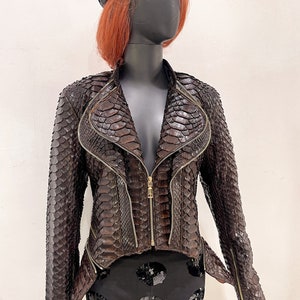 以下が含まれることがあります： ジッパー留めとハイネックのブラウンのスネークスキンレザーのジャケット。ジャケットはフィットしたシルエットと短い丈です。ジャケットは赤い髪と黒いベレー帽をかぶったマネキンが着用しています。