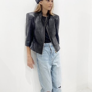 May include: A woman wearing a black snakeskin jacket with a gold zip, light blue jeans with rips, and a black beret.