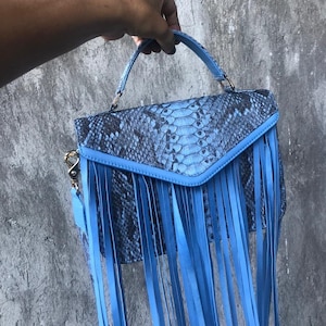 May include: A light blue handbag with a snakeskin pattern and long fringe details. The bag has a top handle and a shoulder strap.