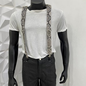 May include: A pair of suspenders with a snake skin pattern. The suspenders are made of leather and have silver buckles. The suspenders are being worn by a mannequin.