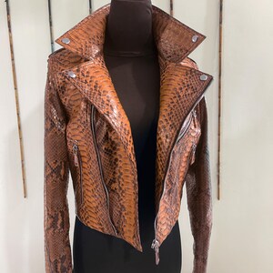 以下が含まれることがあります： ヘビ柄の茶色の革ジャン。ジッパー留めと襟がある。