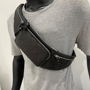 May include: A black leather fanny pack with a textured, embossed design. The bag has two zippered compartments and a silver zipper pull.