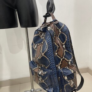 Op de afbeelding: Een blauwe en bruine slangleren rugzak met een ritsvak aan de voorkant. De rugzak heeft een bovenste handvat en verstelbare banden.