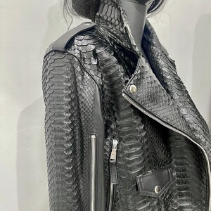 K&ouml;nnte beinhalten: Schwarze Lederjacke mit Schlangenhautmuster. Die Jacke hat einen Rei&szlig;verschluss und zwei Taschen.